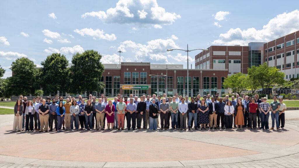Large group of Oak Ridge Leadership Computing Facility Summit participants posing for a photo outdoors at Oak Ridge National Laboratory with laboratory buildings in the background.