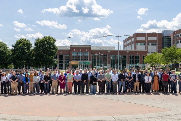 Large group of Oak Ridge Leadership Computing Facility Summit participants posing for a photo outdoors at Oak Ridge National Laboratory with laboratory buildings in the background.